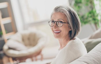 An older woman with gray hair and glasses sits on a couch, smiling at the camera. She is wearing a white sweater. The background is softly lit with some plants visible.